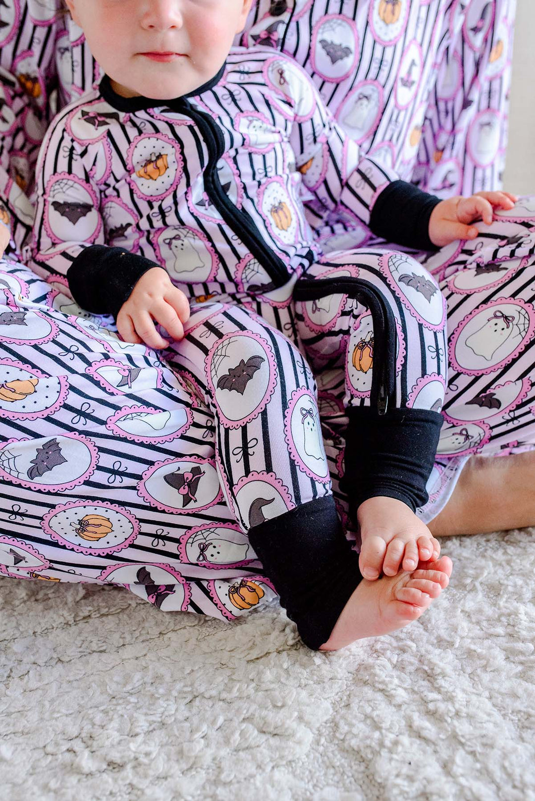 Baby sitting in an adult's lap, both wearing matching Caden Lane pink and black striped Halloween pajamas with ghost, bat, and pumpkin cameo print, the baby's romper with black cuffs and a black bow.