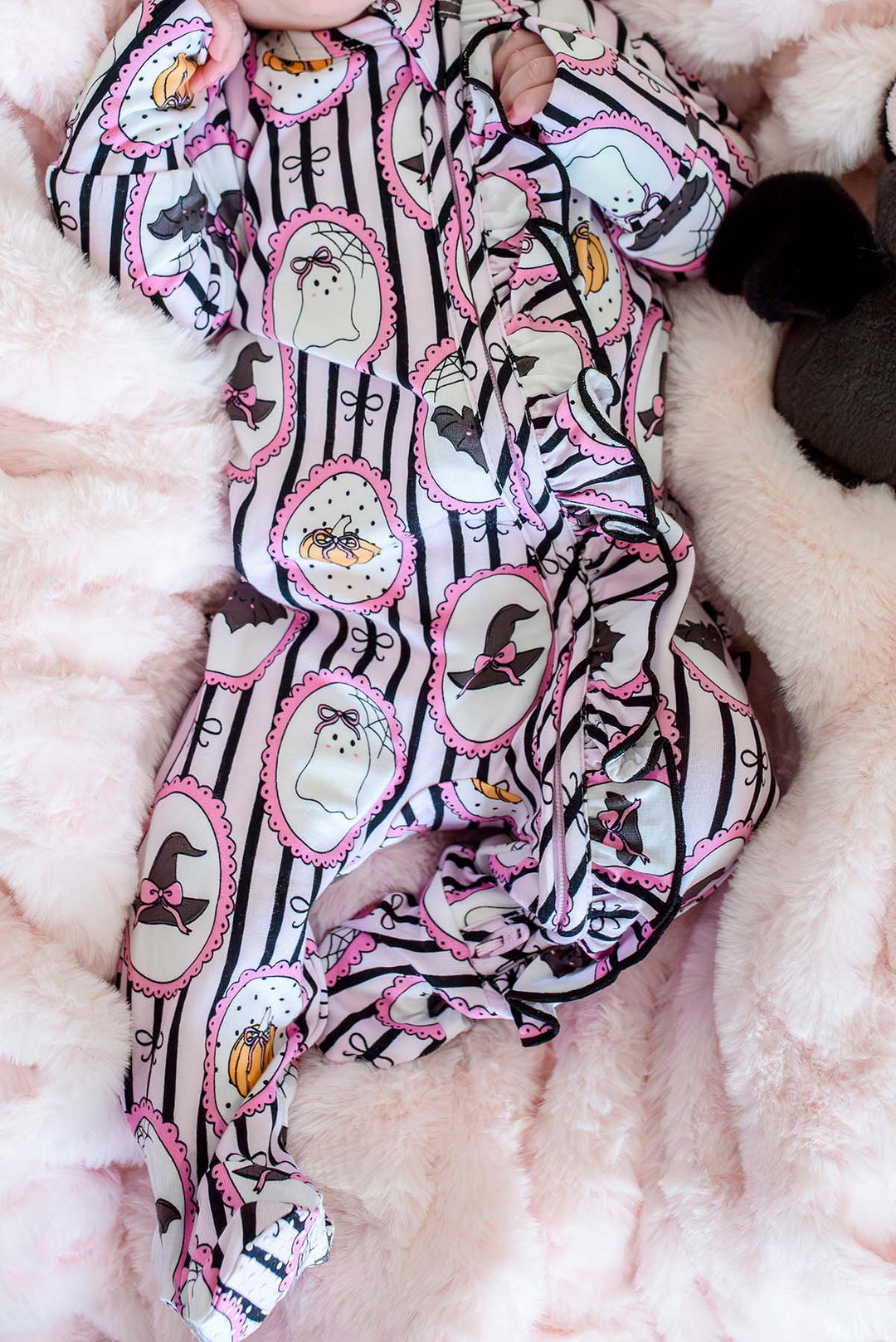 Close-up of a baby lying on a pink plush blanket, wearing a Halloween-print footed sleeper with ghost, witch hat, and pumpkin designs.