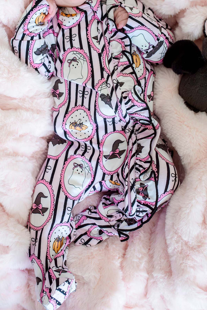 Close-up of a baby lying on a pink plush blanket, wearing a Halloween-print footed sleeper with ghost, witch hat, and pumpkin designs.