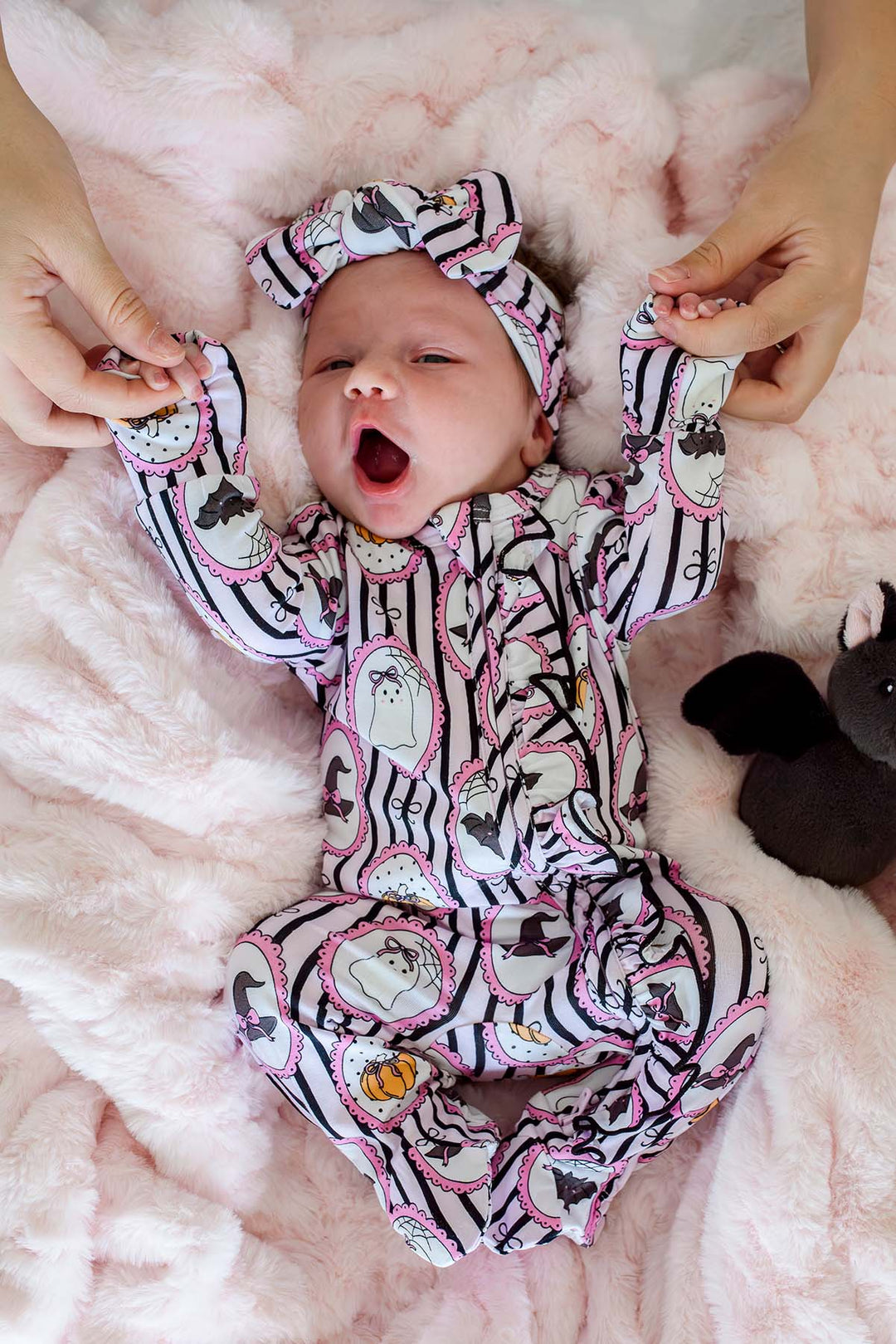 A baby wearing a Halloween-themed footed sleeper and matching headband with black and white stripes, pink accents, and ghost, bat, and pumpkin designs, lying on a pink plush blanket while adult hands hold the baby's hands.