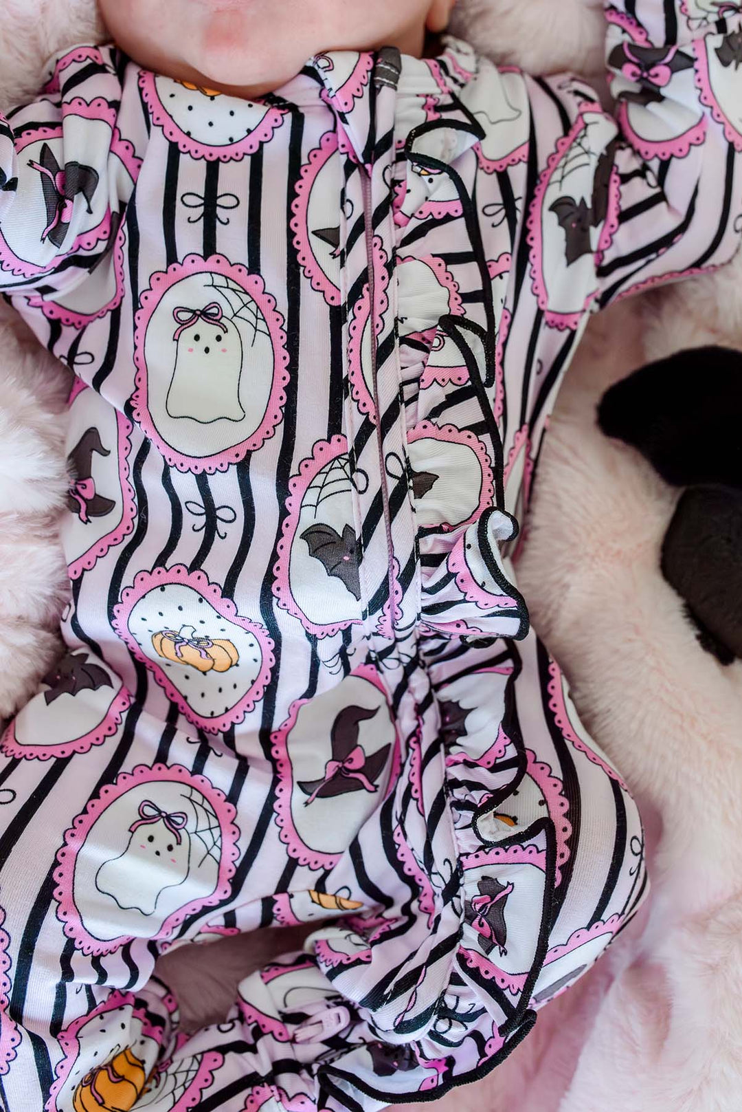 A close-up of a baby's torso wearing a Halloween-themed footed sleeper. The sleeper has a white base with black vertical stripes, pink scalloped borders, and cartoon ghost, bat, spider, and pumpkin motifs.