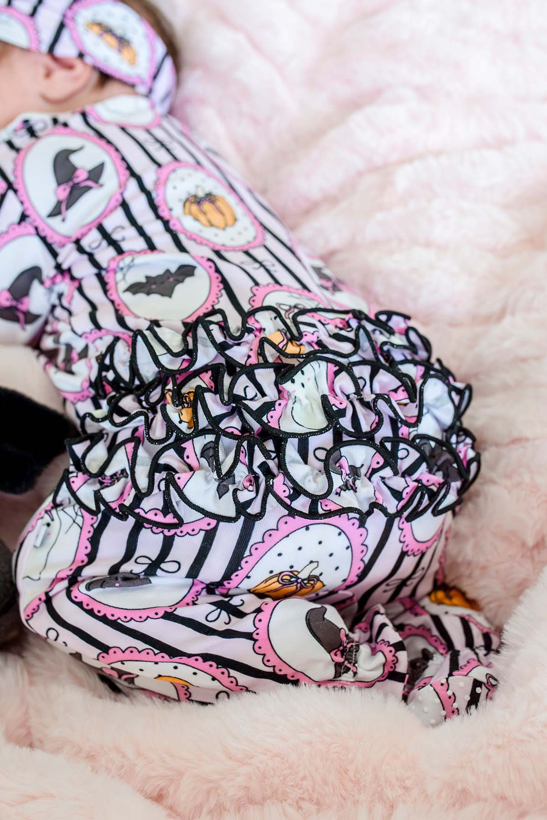 A baby wearing a Halloween-themed bodysuit and matching hat, lying on a soft pink blanket. The bodysuit has a black and white striped pattern with pink ruffles, featuring cartoon ghosts, bats, and pumpkins.