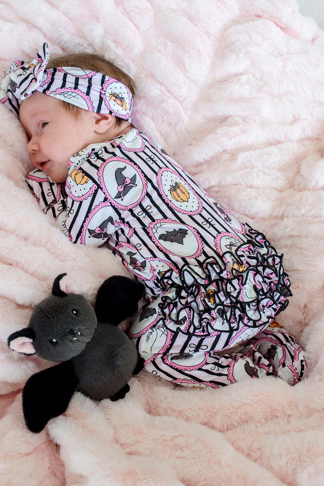 Newborn wearing a Caden Lane pink and black striped Halloween ruffle footie with ghost, pumpkin, and witch hat frame print, plus a matching bow headband, lying next to a plush black bat toy