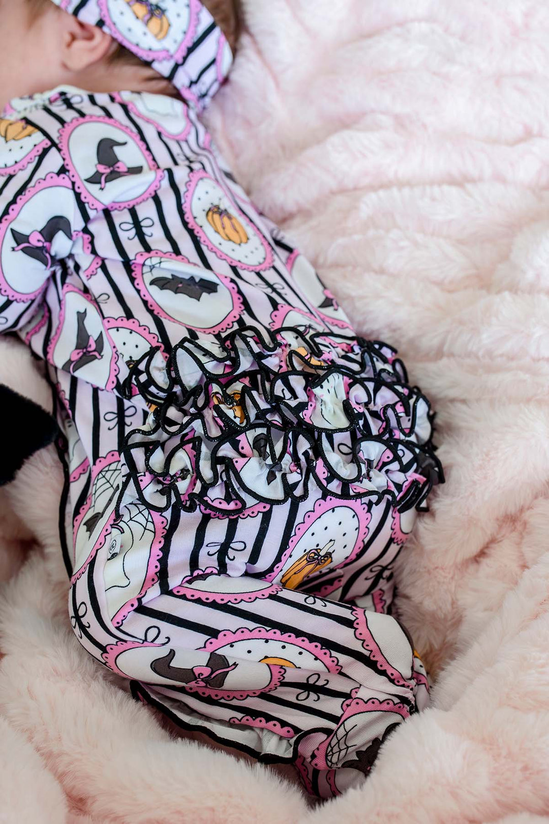 A baby lying on a pink plush blanket, wearing a Halloween-print footed sleeper and matching headband with witch hat, bat, ghost, and pumpkin designs.
