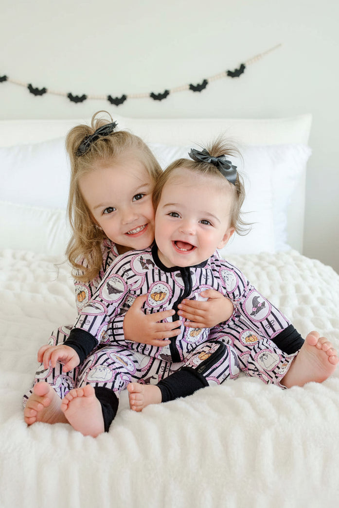 Baby and toddler sitting both wearing matching Caden Lane pink and black striped Halloween pajamas with ghost, bat, and pumpkin cameo print, the baby's romper with black cuffs and a black bows.