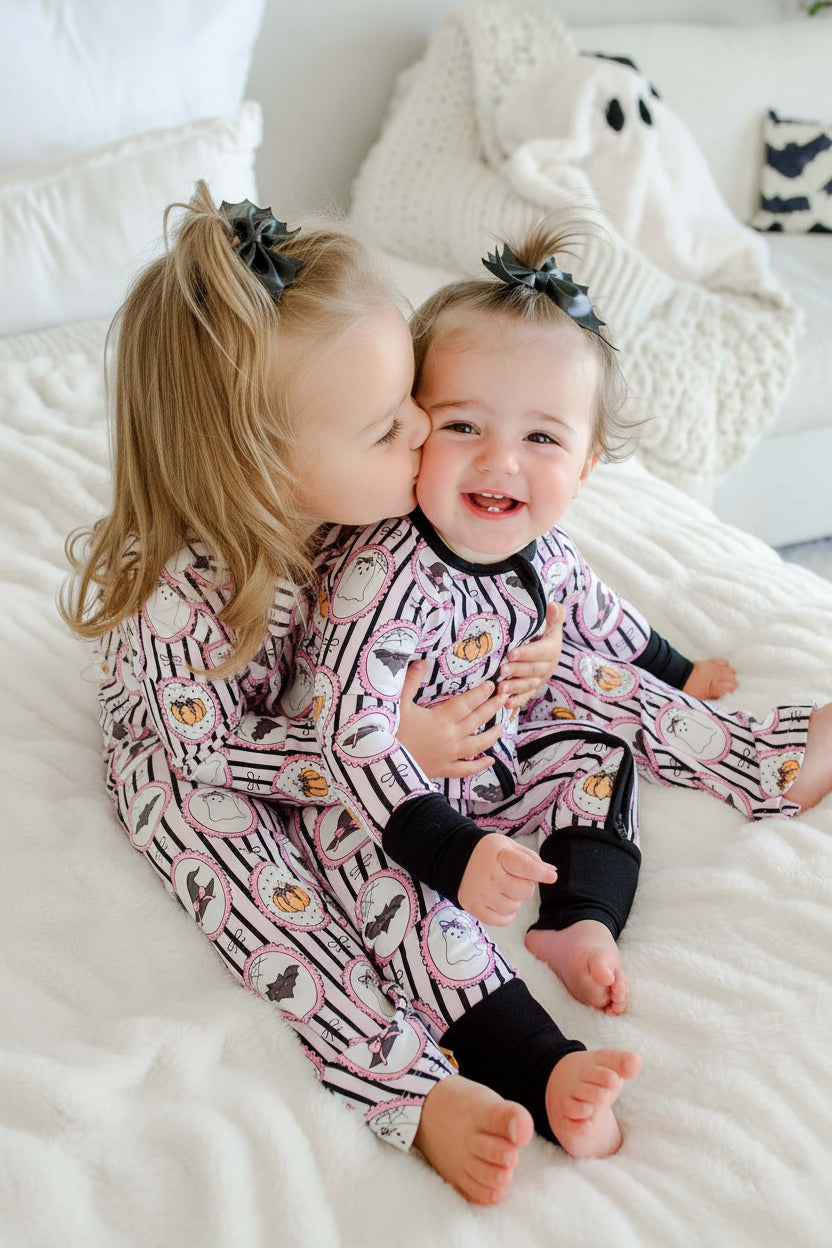 Baby and toddler sitting both wearing matching Caden Lane pink and black striped Halloween pajamas with ghost, bat, and pumpkin cameo print, the baby's romper with black cuffs and a black bows.