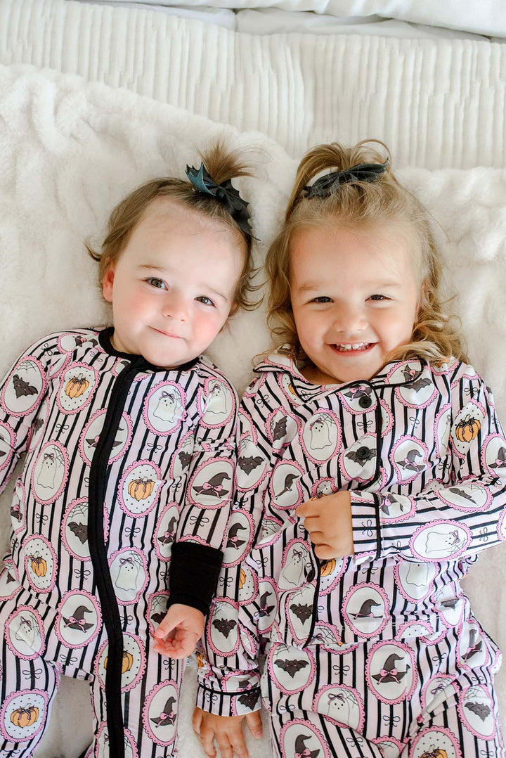 Baby and toddler laying both wearing matching Caden Lane pink and black striped Halloween pajamas with ghost, bat, and pumpkin cameo print, the baby's romper with black cuffs and a black bows.