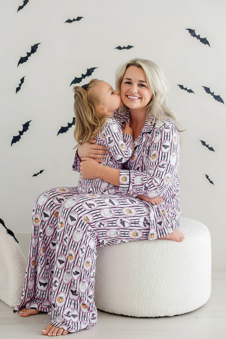 A woman and a young girl wearing matching purple and white patterned pajamas sit together on a round white pouf, with black bat decals on a light wall behind them.
