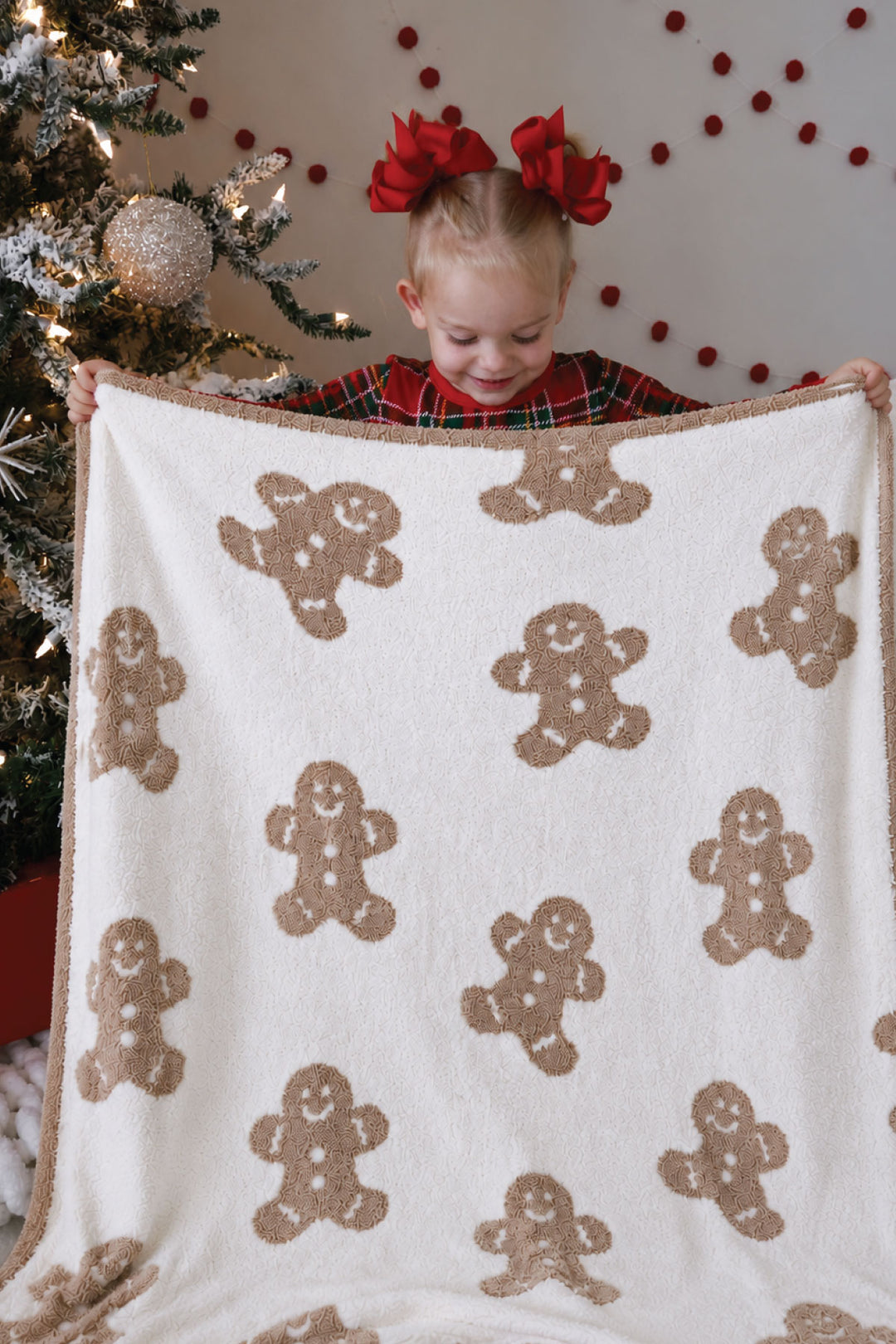 Toddler girl in red plaid pajamas holding up a Caden Lane cream sherpa blanket with an allover taupe gingerbread man print, standing beside a Christmas tree