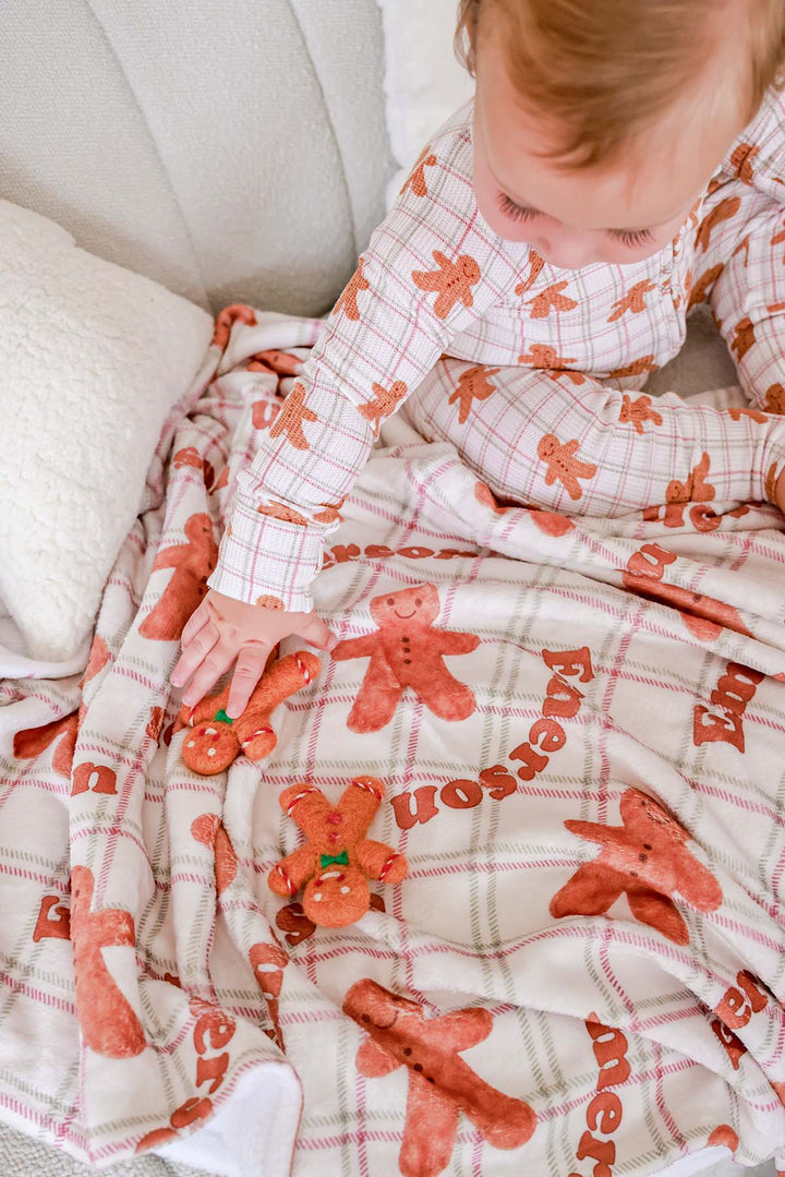 Caden Lane toddler wearing soft bamboo gingerbread cookie print Christmas pajamas while playing with matching gingerbread plush toys on a coordinating holiday blanket, creating a cozy festive scene perfect for Christmas morning, holiday gifting, and family traditions.