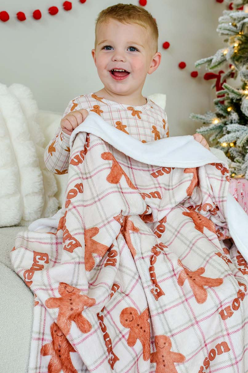toddler sitting by a christmas tree holding a personalized Caden Lane gingerbread print kids christmas blanket