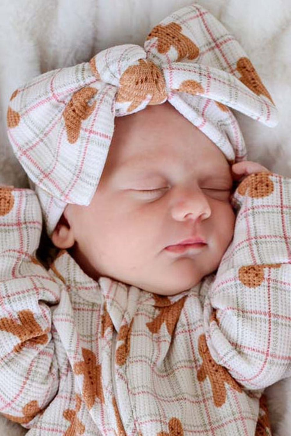 A sleeping newborn baby wearing a Caden Lane gingerbread-themed footed sleeper and matching headband, lying on a white textured blanket next to a small gingerbread doll.