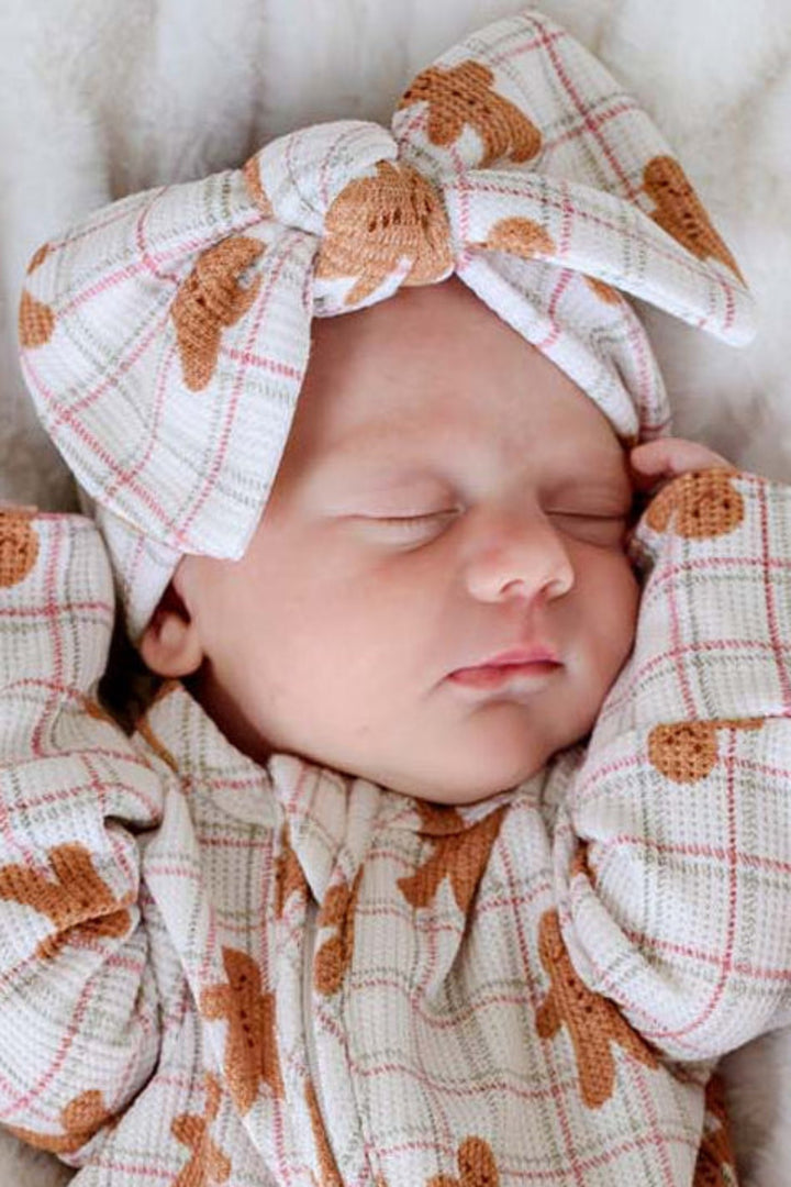 A sleeping newborn baby wearing a Caden Lane gingerbread-themed footed sleeper and matching headband, lying on a white textured blanket next to a small gingerbread doll.