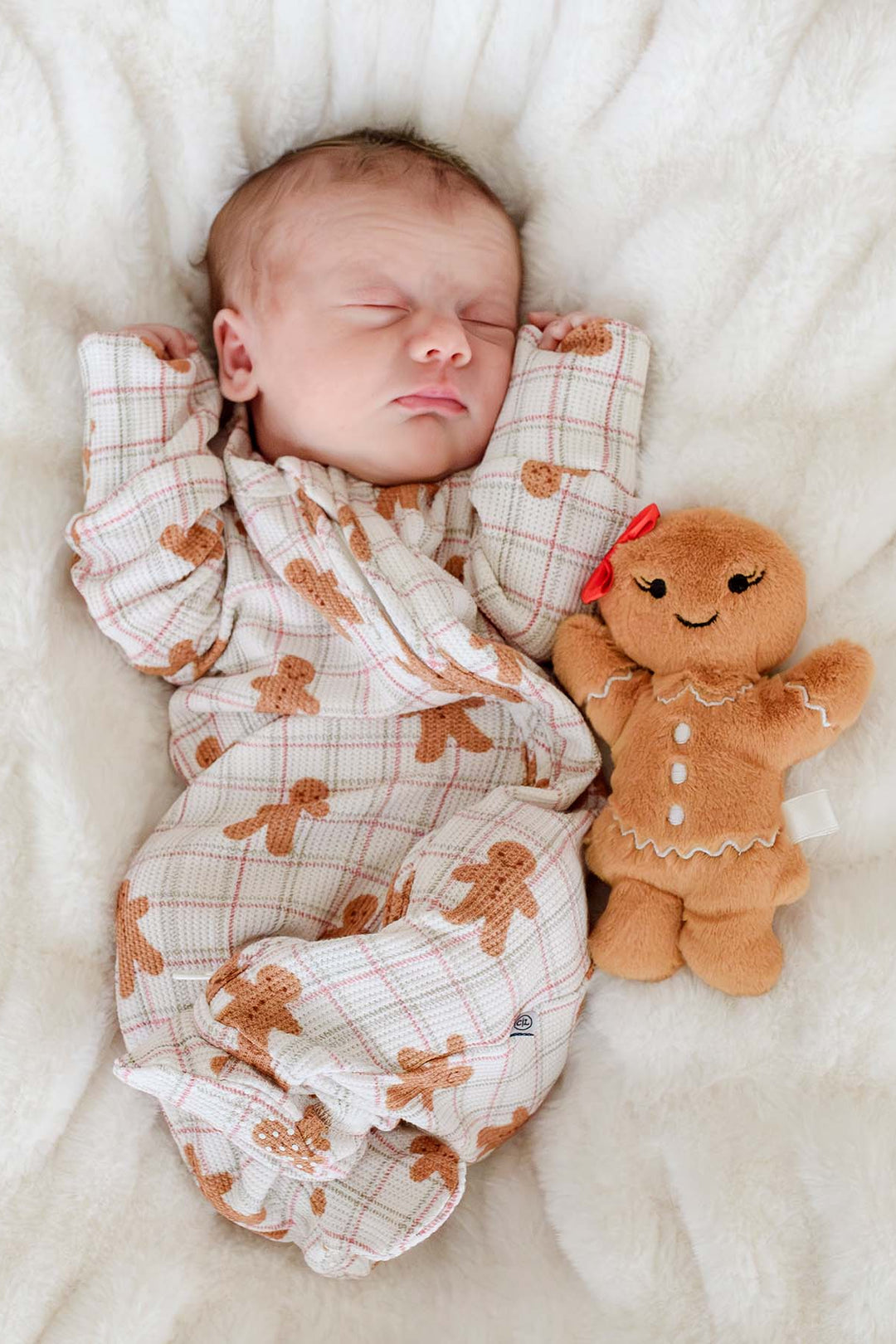 A sleeping newborn baby is swaddled in a soft cream and brown gingerbread print blanket and lies on a white textured blanket, flanked by wrapped Christmas presents and a small gingerbread doll.