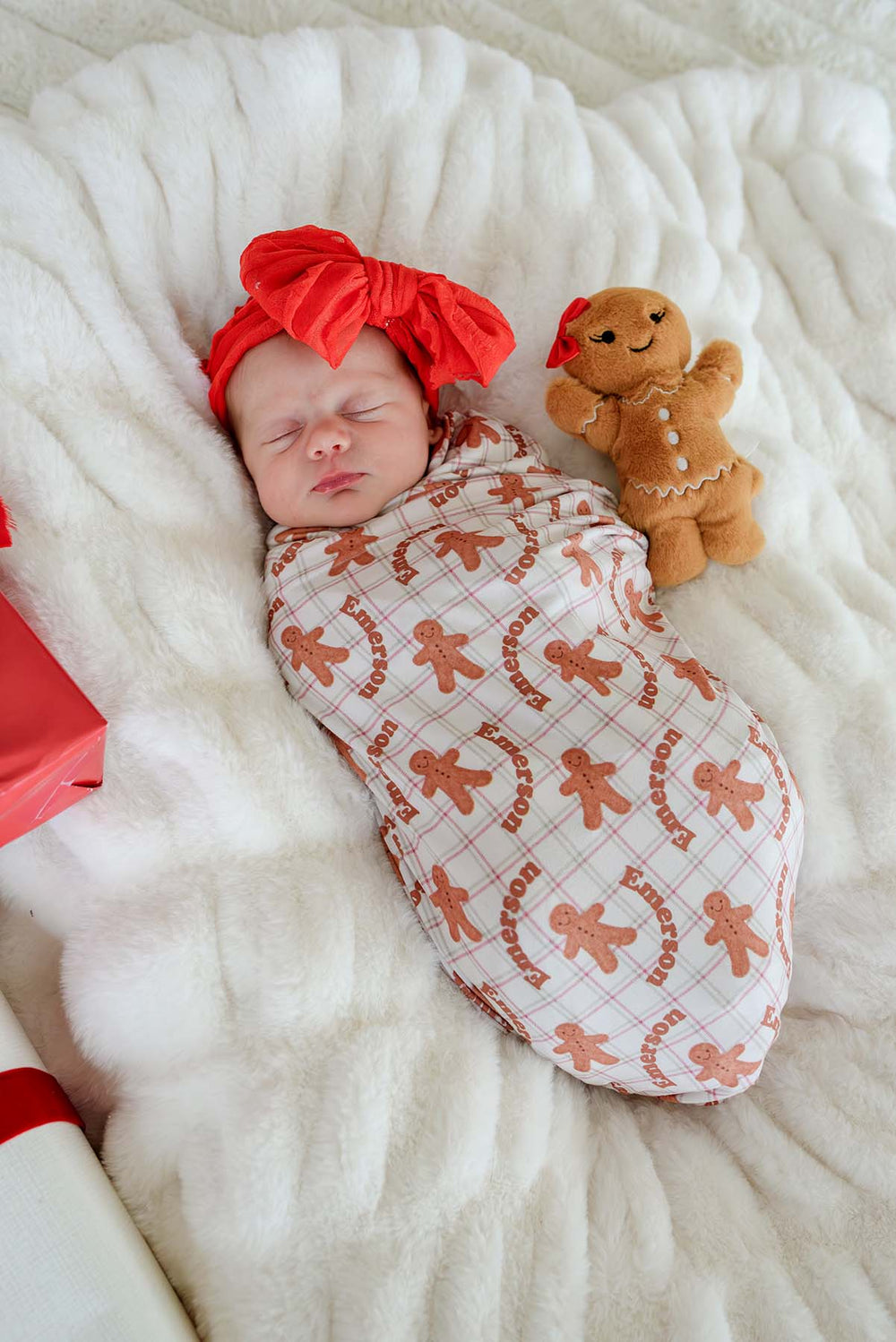 Caden Lane personalized bamboo Christmas swaddle blanket in a gingerbread cookie print with the name "Emerson," wrapped around a sleeping newborn beside a gingerbread plush, creating a cozy holiday keepsake for baby's first Christmas and seasonal gifting.