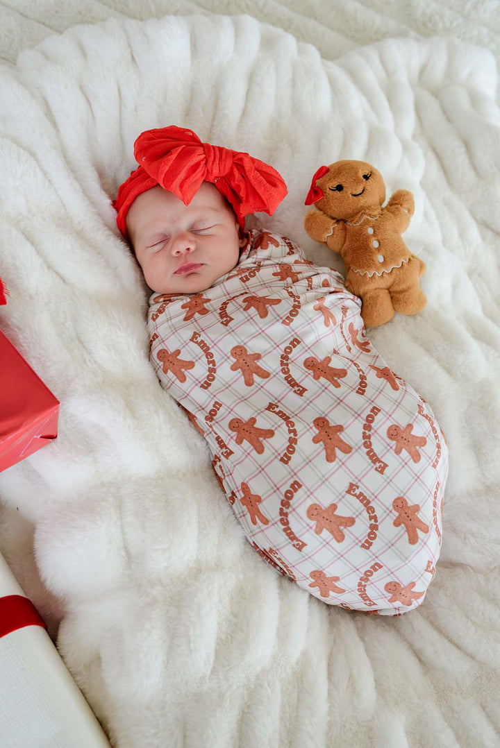 Caden Lane personalized bamboo Christmas swaddle blanket in a gingerbread cookie print with the name "Emerson," wrapped around a sleeping newborn beside a gingerbread plush, creating a cozy holiday keepsake for baby's first Christmas and seasonal gifting.