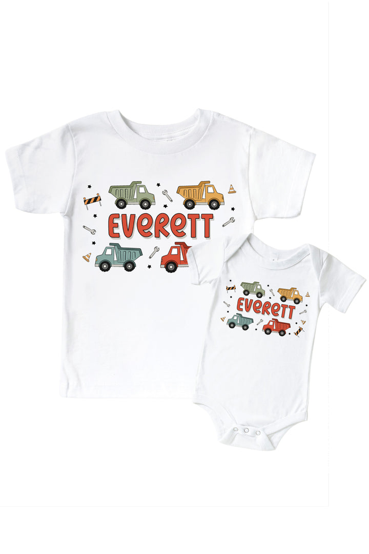 Flat lay of a Caden Lane white tee in a Busy Builder print featuring four colorful dump trucks, reading "Everett," paired with a matching personalized bodysuit.
