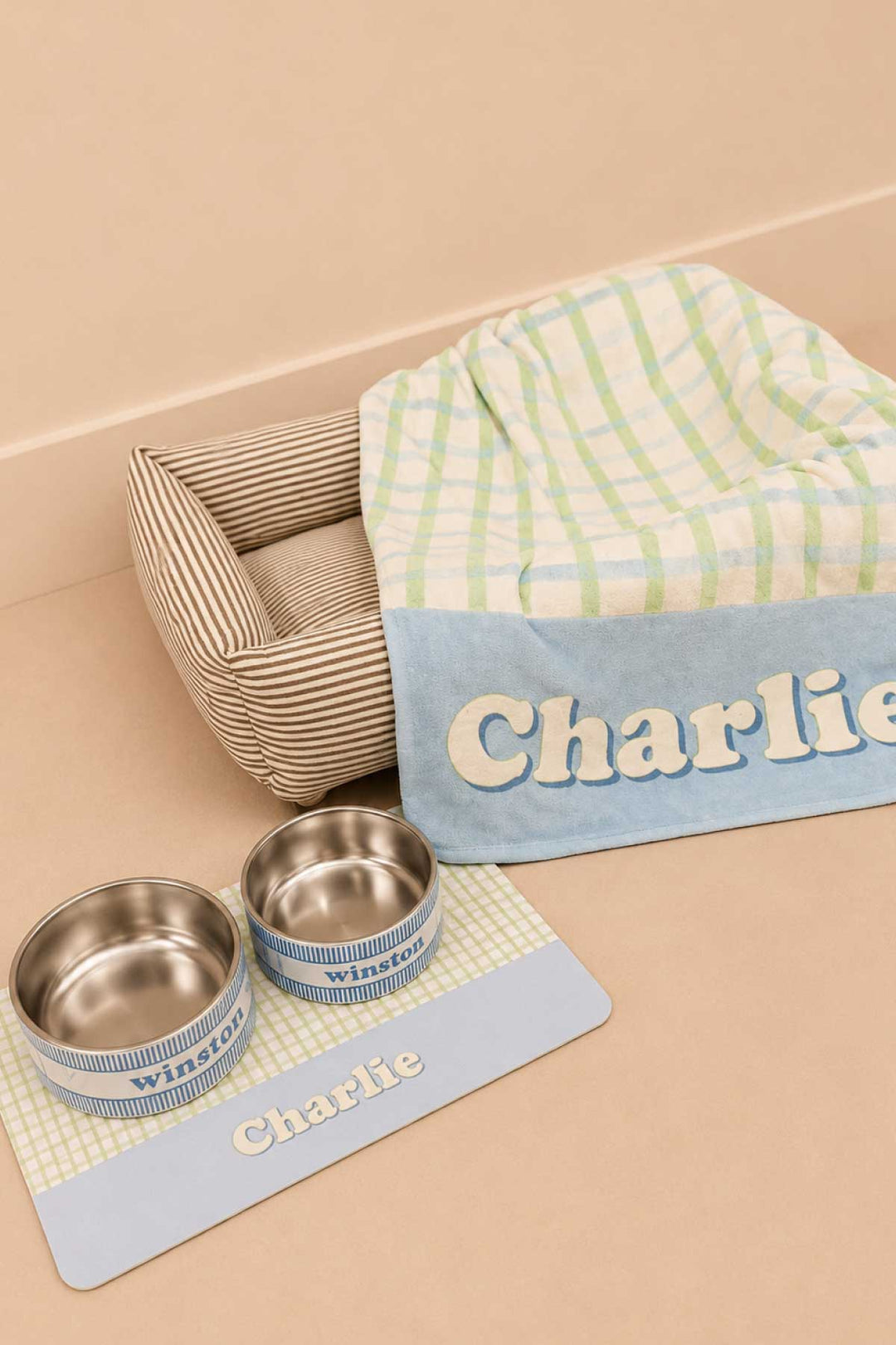 Lifestyle photo showing a picnic plaid pet blanket draped over a wooden pet bed frame with a personalized placemat labeled "Charlie" and matching double bowls labeled "Winston" in front