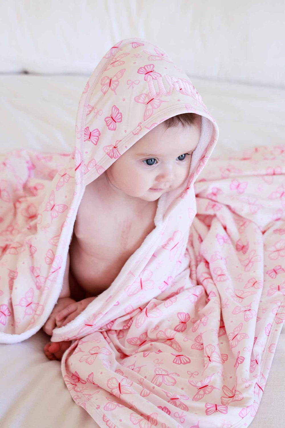 Embroidered Bamboo Hooded Towel in Blooming Butterflies print - baby sitting on white surface wrapped in the pink bamboo towel, with hood on and butterfly print visible
