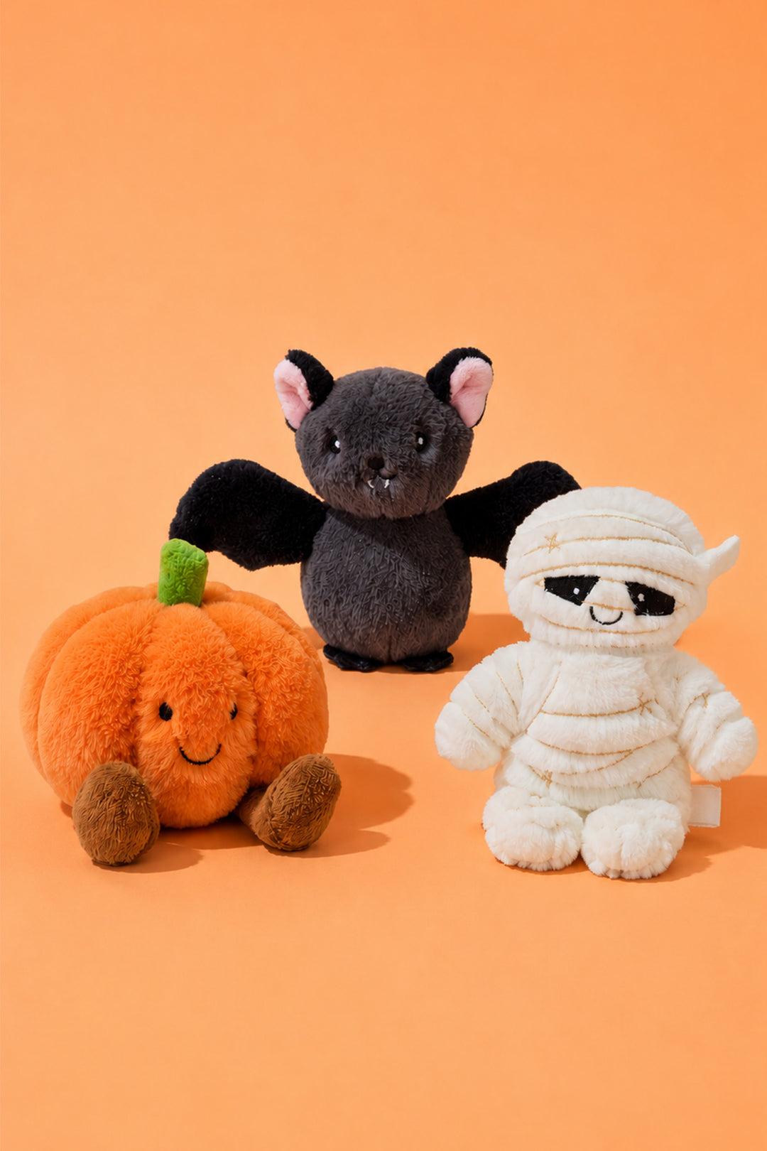 Caden Lane Halloween plush toys on an orange background: a smiling orange pumpkin with brown feet, a gray bat with black wings and pink-lined ears standing in the center, and a white mummy plush with gold stitching and a black-masked face sitting on the right.