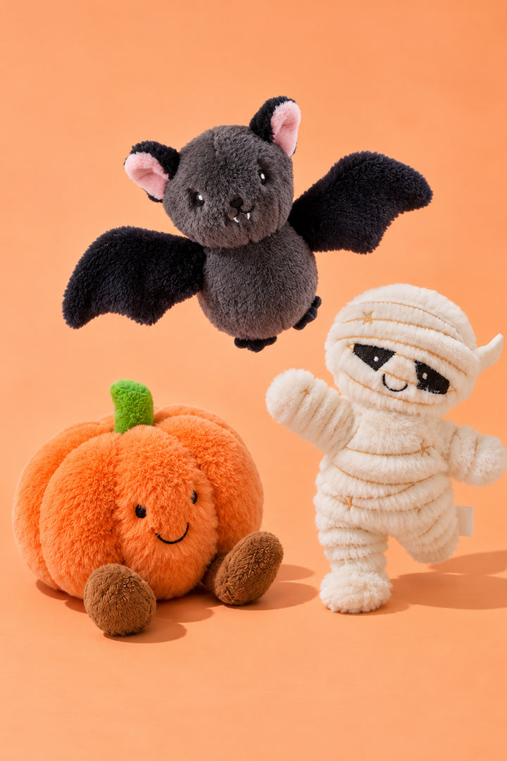Caden Lane's gray bat plush flies above a smiling orange pumpkin plush and a white mummy plush, who raises one arm in a wave, against an orange background.