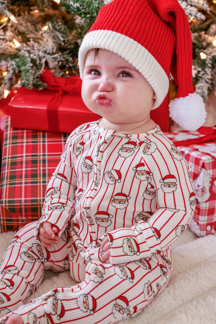 Baby wearing a Caden Lane Holly & Jolly  print bamboo waffle convertible zipper romper featuring a vintage Santa print and red stripes, paired with a red Santa hat while sitting in front of a decorated Christmas tree and wrapped holiday gifts.