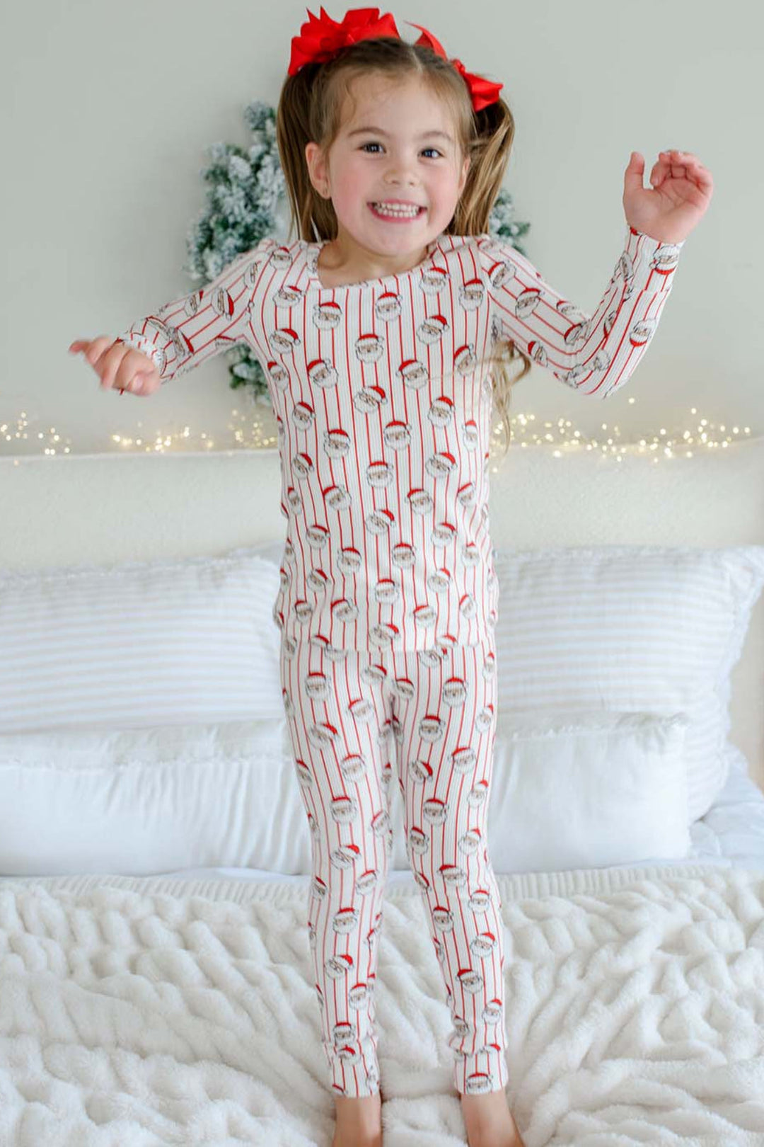 Caden Lane girls' bamboo Christmas pajamas with a cheerful Santa stripe print, featuring a toddler jumping on the bed in cozy, festive holiday sleepwear perfect for Christmas morning.