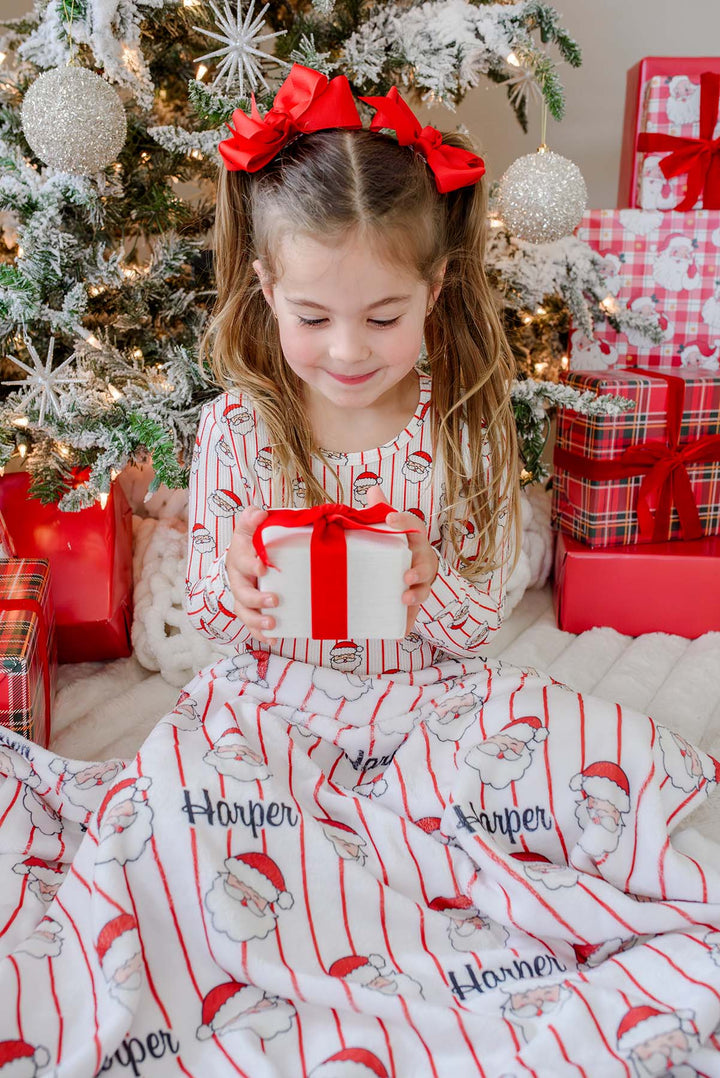 Caden Lane personalized Santa stripe Christmas blanket with custom name, paired with matching bamboo pajamas as a child opens a holiday gift by the Christmas tree.