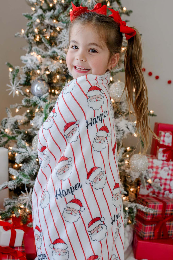 Caden Lane personalized Christmas blanket featuring a festive Santa stripe print with custom name, wrapped around a smiling child by the Christmas tree for cozy holiday memories.