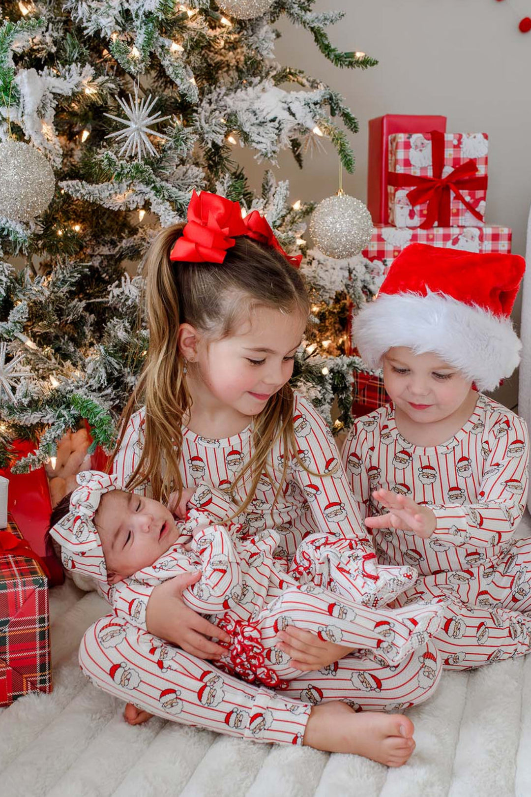 Caden Lane matching bamboo Christmas pajamas featuring a festive Santa stripe print, worn by siblings and a newborn in coordinating holiday sleepwear while gathered by the Christmas tree for a cozy family Christmas photo.
