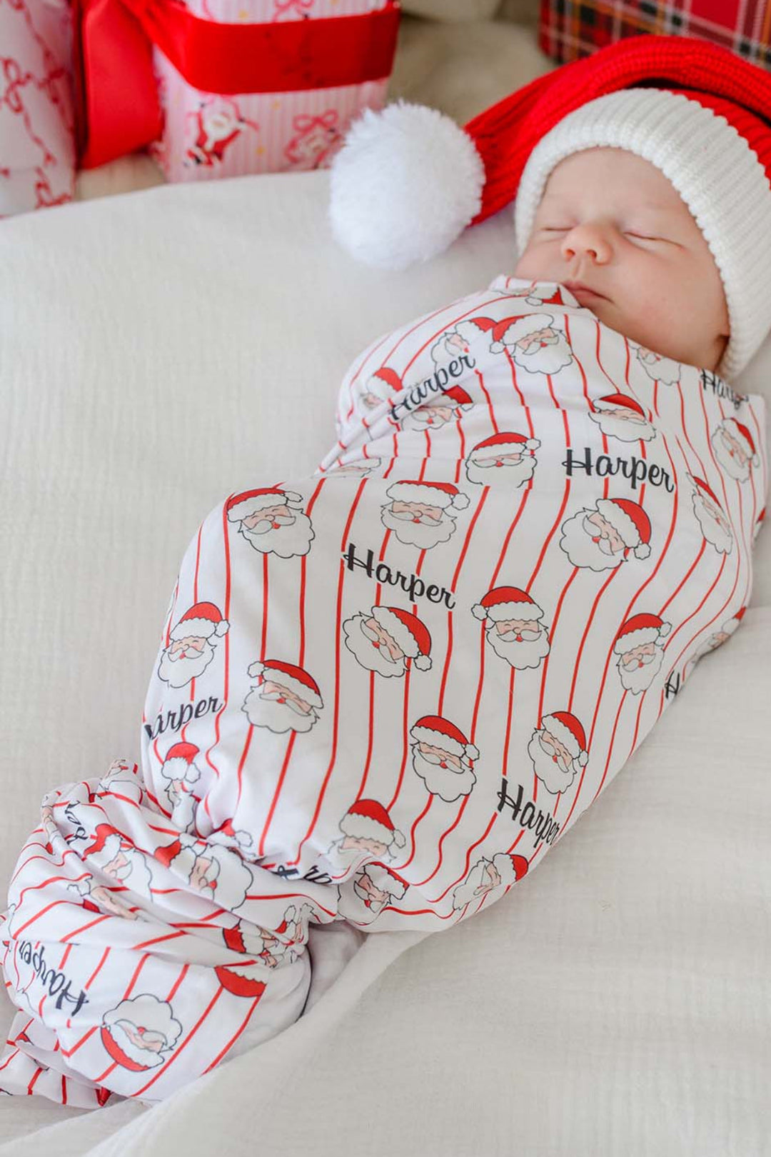 Personalized Holly & Jolly Christmas bamboo swaddle blanket with Santa print and red pinstripes wrapped around a sleeping newborn wearing a festive Santa hat.
