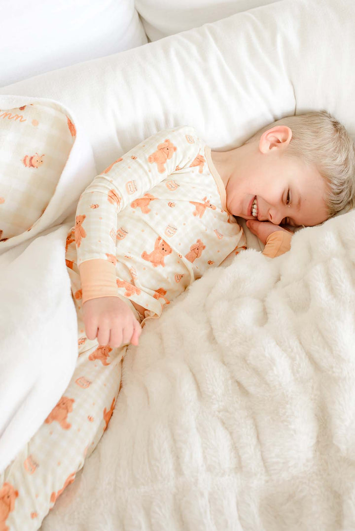 Laughing toddler boy wearing a Caden Lane honey bear print two-piece pajama set, laying on a bed