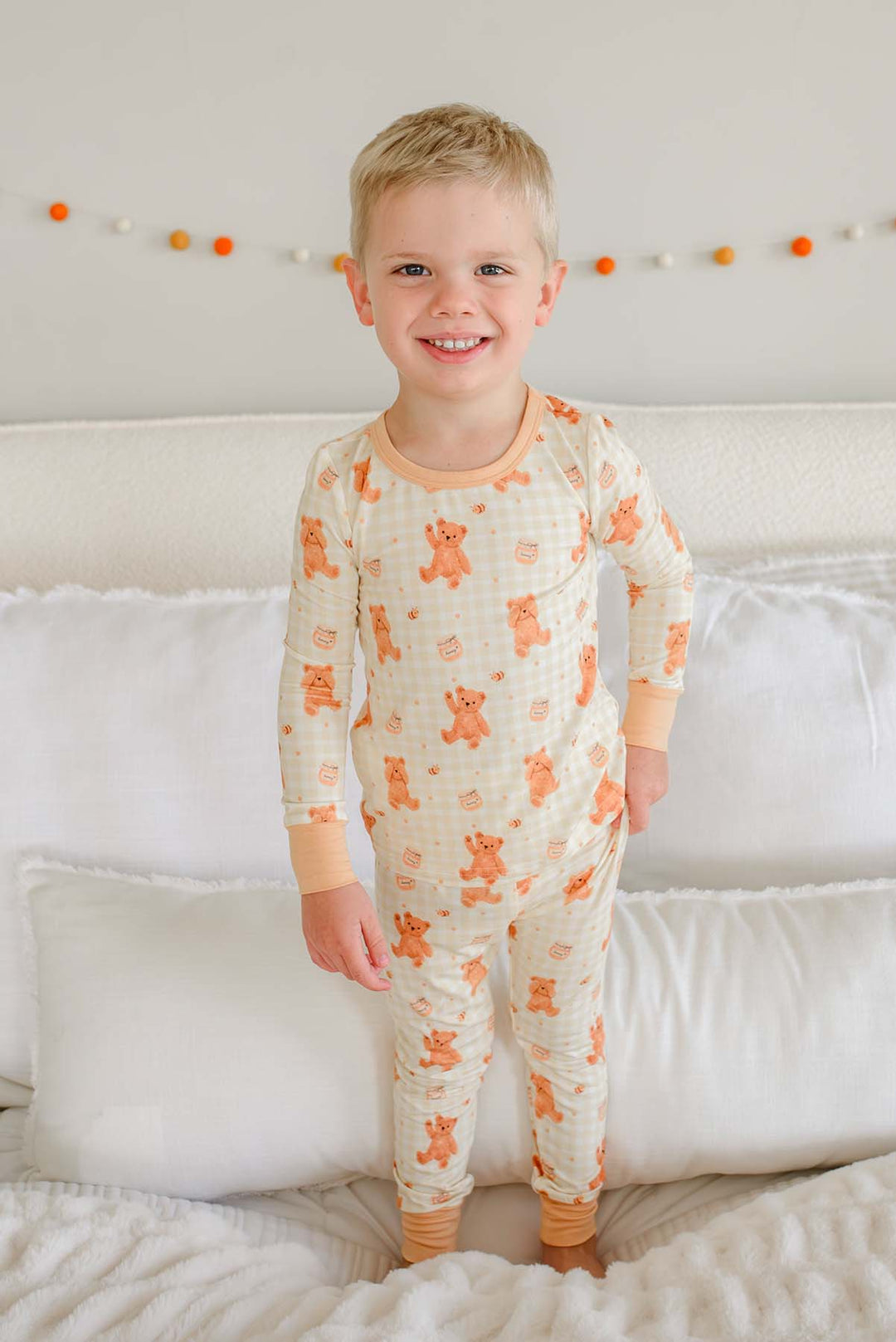 Laughing toddler boy wearing a Caden Lane honey bear print two-piece pajama set, standing on a bed