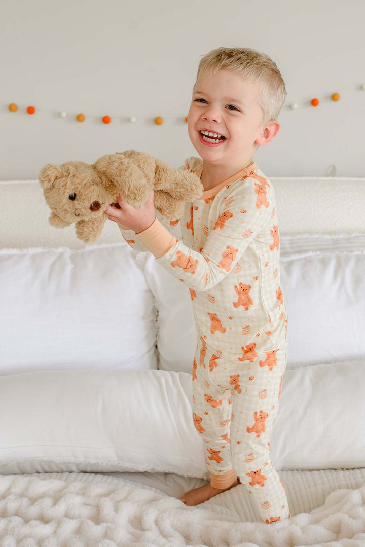 Laughing toddler boy wearing a Caden Lane honey bear print two-piece pajama set, standing on a bed holding up a plush teddy bear
