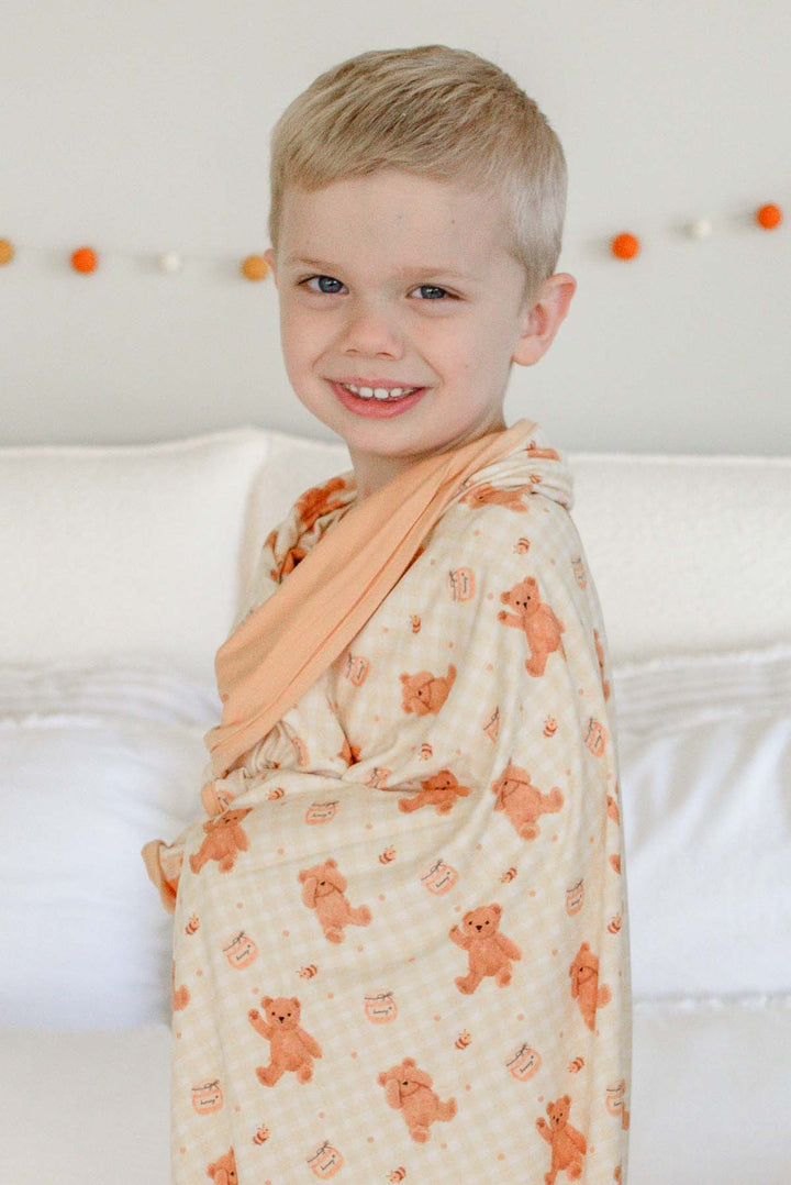 Smiling boy wearing Caden Lane honey bear print pajamas, wrapped in a matching Caden Lane bamboo blanket while standing on a bed