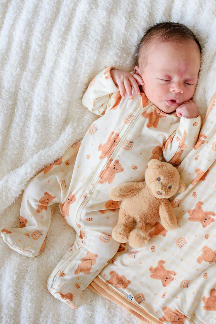 A sleeping newborn baby wearing a cream-colored footed sleeper with a light orange teddy bear pattern, lying on a white blanket next to a matching baby blanket and a small teddy bear toy.