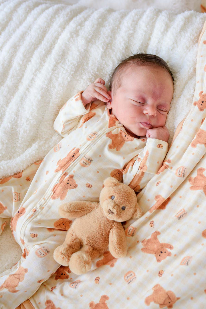 A sleeping newborn baby wearing a cream-colored zip-up onesie with a brown teddy bear pattern, lying on a white textured blanket next to a small plush teddy bear.