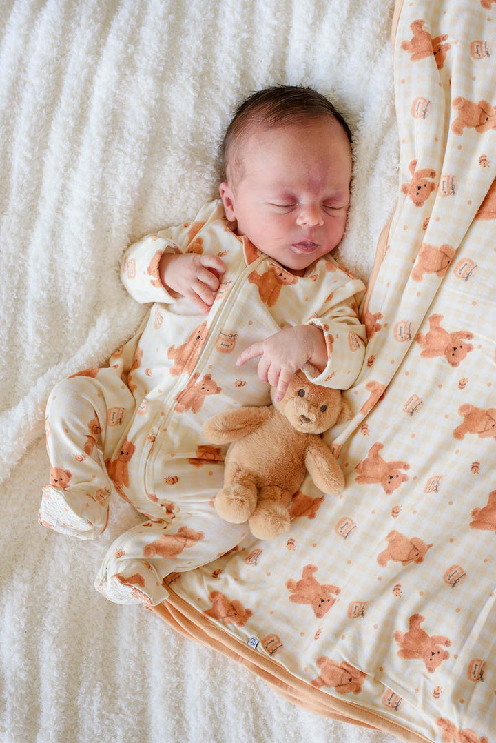 Newborn baby sleeping in a Caden Lane honey bear print footie pajama, lying on a fluffy white blanket next to a matching Caden Lane plush teddy bear and honey bear blanket