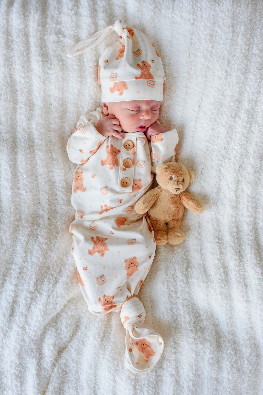Sleeping newborn wearing a Caden Lane honey bear print knotted gown and matching sleeper cap, lying on a fluffy white blanket next to a plush teddy bear