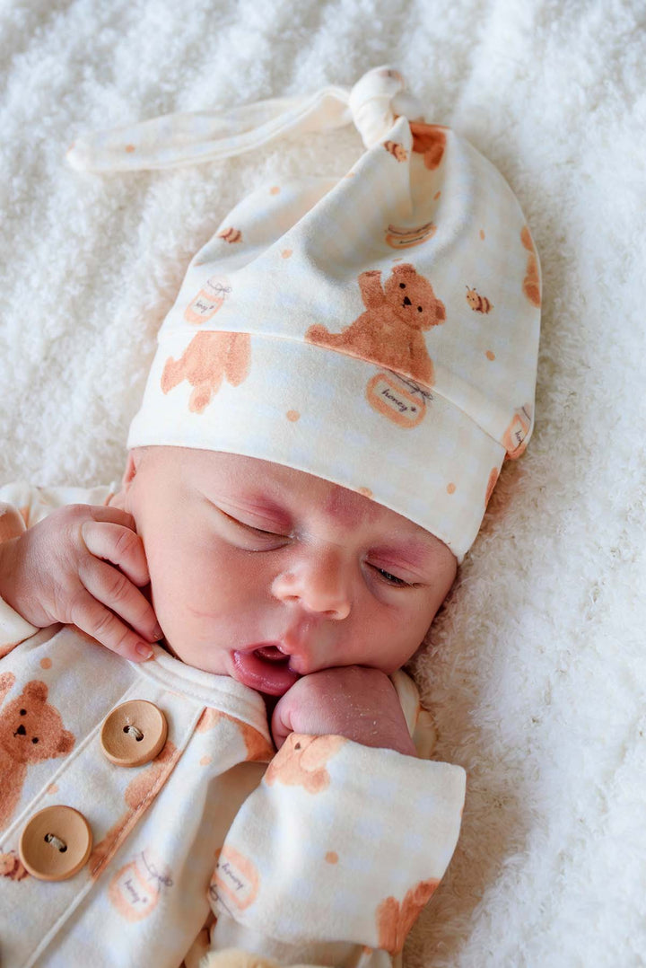 Newborn wearing a Caden Lane honey bear print knotted gown and matching sleeper cap, lying on a fluffy white blanket