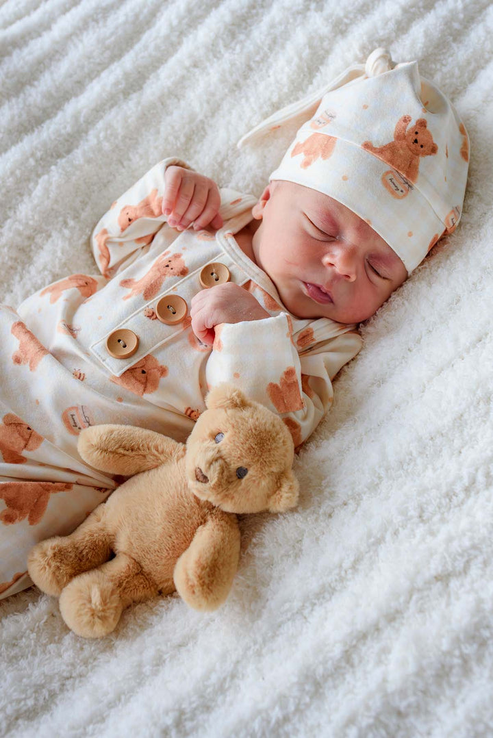Sleeping newborn wearing a Caden Lane honey bear print knotted gown and matching sleeper cap, lying on a fluffy white blanket next to a plush teddy bear