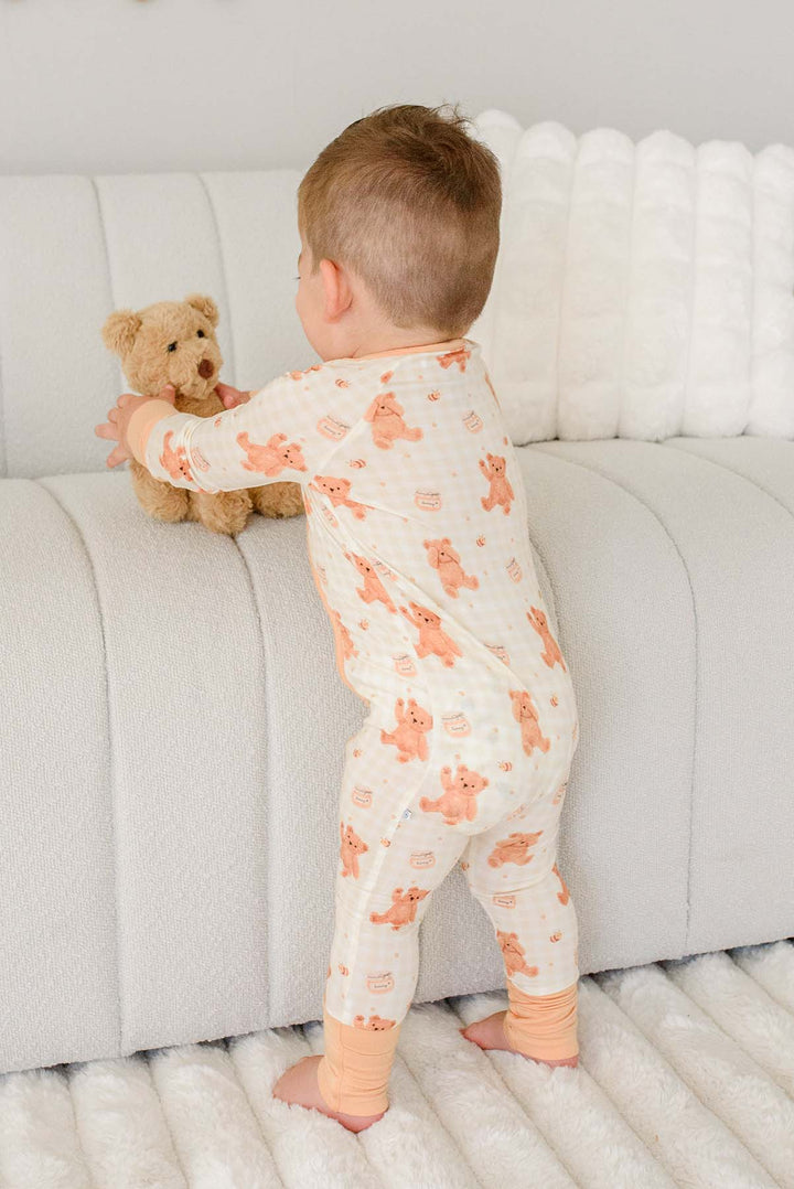Toddler standing barefoot near a couch wearing a Caden Lane honey bear print convertible romper, with a plush teddy bear