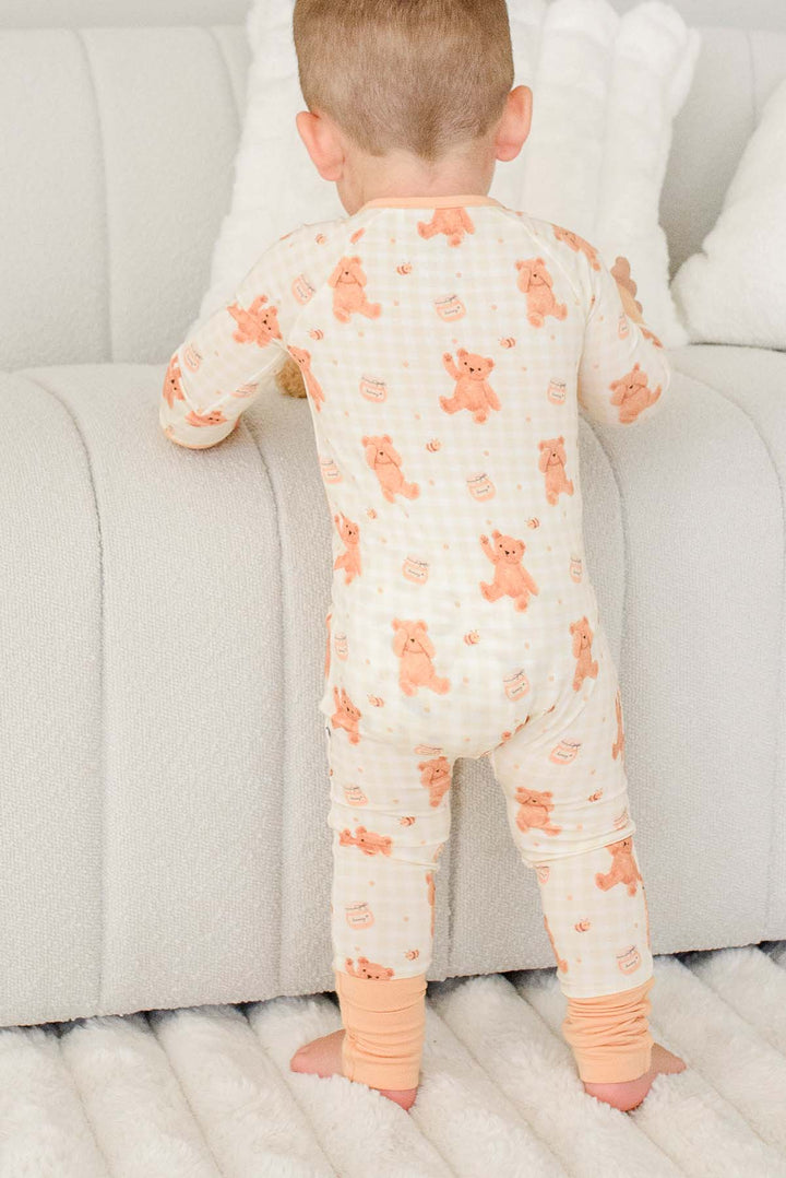 Toddler standing barefoot near a couch wearing a Caden Lane honey bear print convertible romper