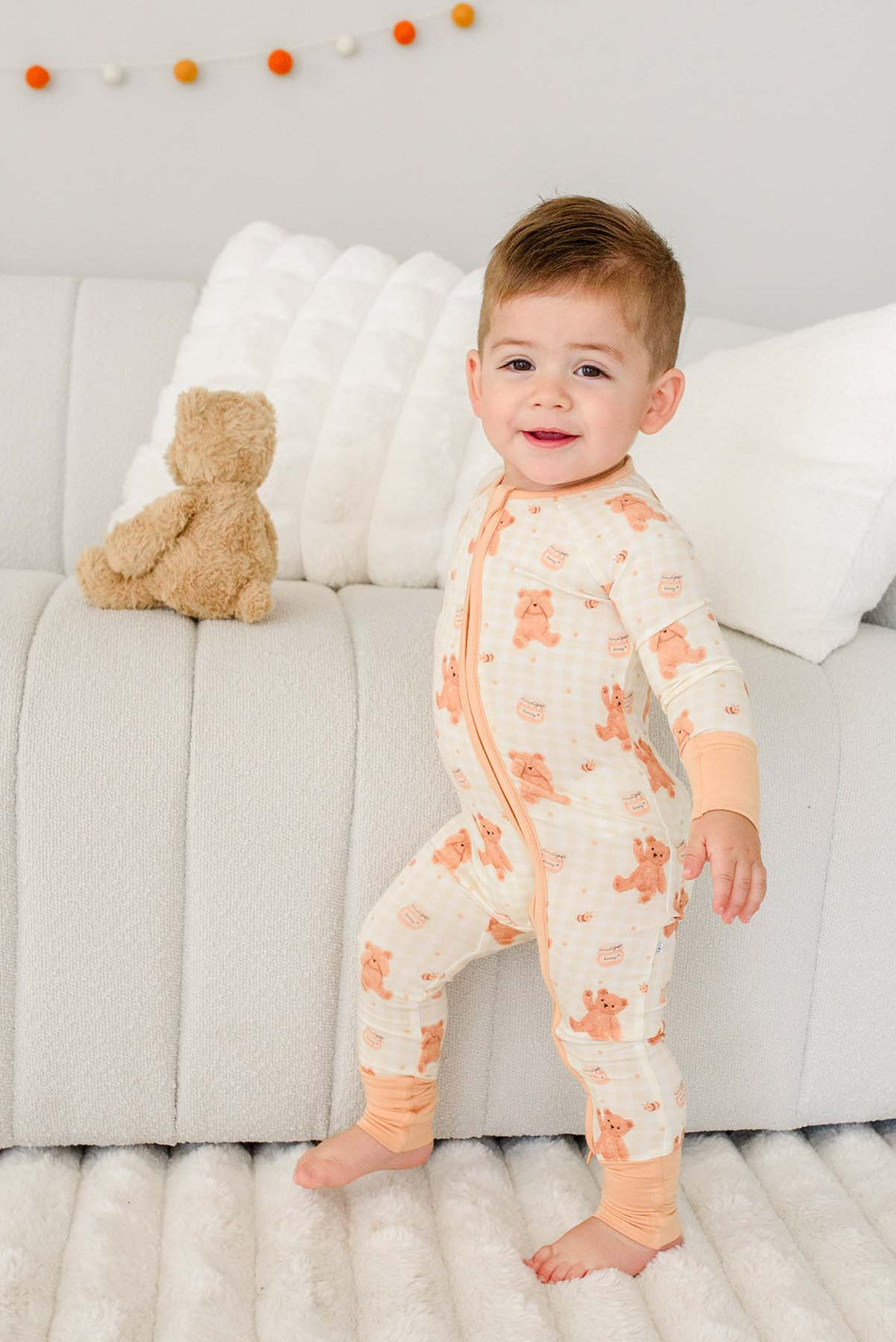Smiling toddler standing barefoot on a couch wearing a Caden Lane honey bear print convertible romper, with a plush teddy bear on the cushion behind him
