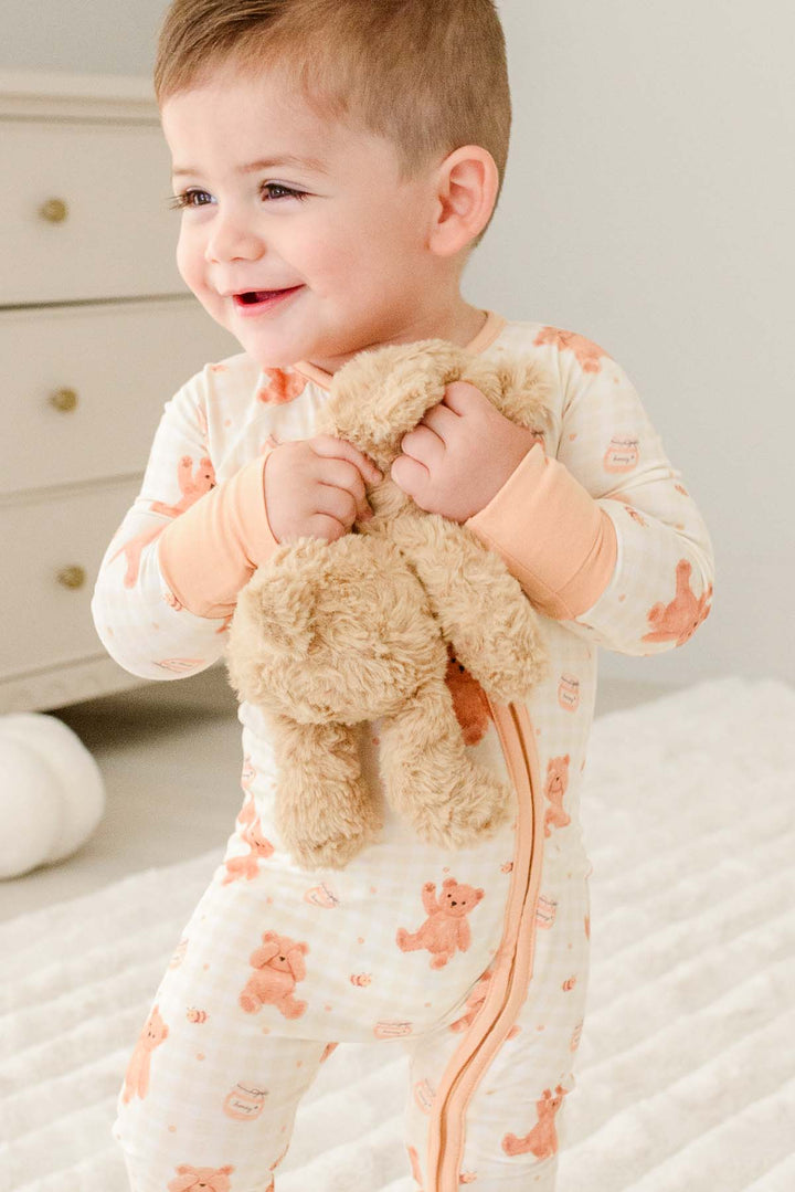 Smiling toddler standing wearing a Caden Lane honey bear print convertible romper, holding a plush teddy bear 