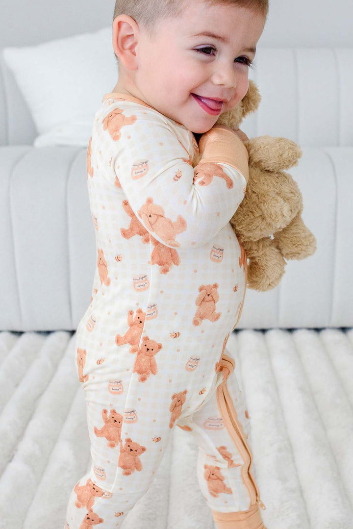 Smiling toddler standing wearing a Caden Lane honey bear print convertible romper, holding a plush teddy bear 