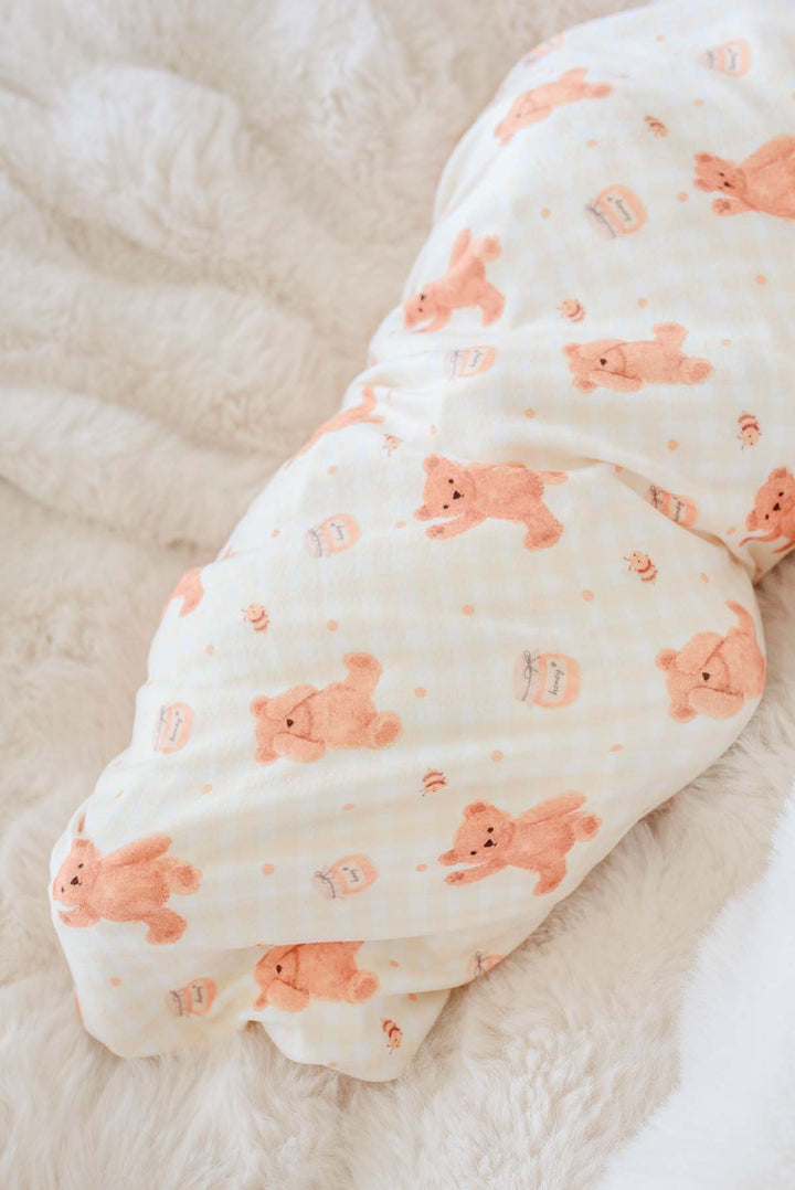 Newborn swaddled in a Caden Lane honey bear print swaddle blanket, lying on a fluffy white blanket