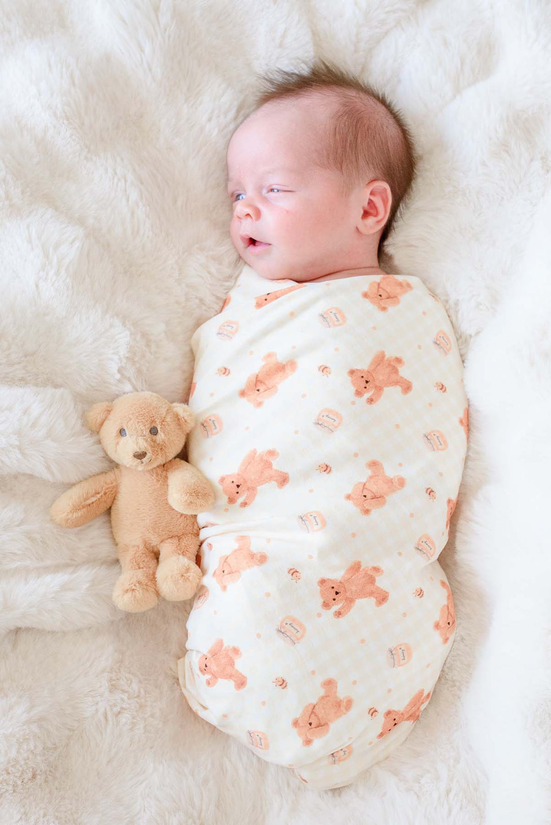 Newborn swaddled in a Caden Lane honey bear print swaddle blanket, lying on a fluffy white blanket next to a plush teddy bear