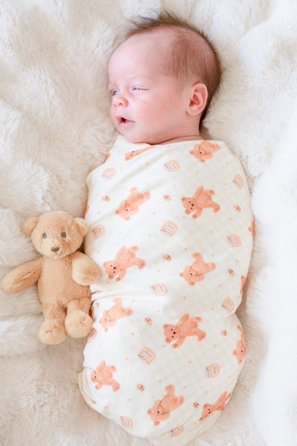Newborn swaddled in a Caden Lane honey bear print swaddle blanket, lying on a fluffy white blanket next to a plush teddy bear