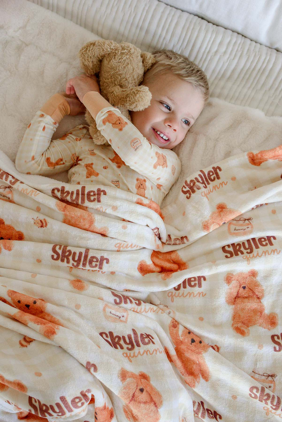 Smiling boy in Caden Lane honey bear print pajamas laying on a bed, holding a plush teddy bear with a personalized Caden Lane blanket printed with 'Skyler Quinn'